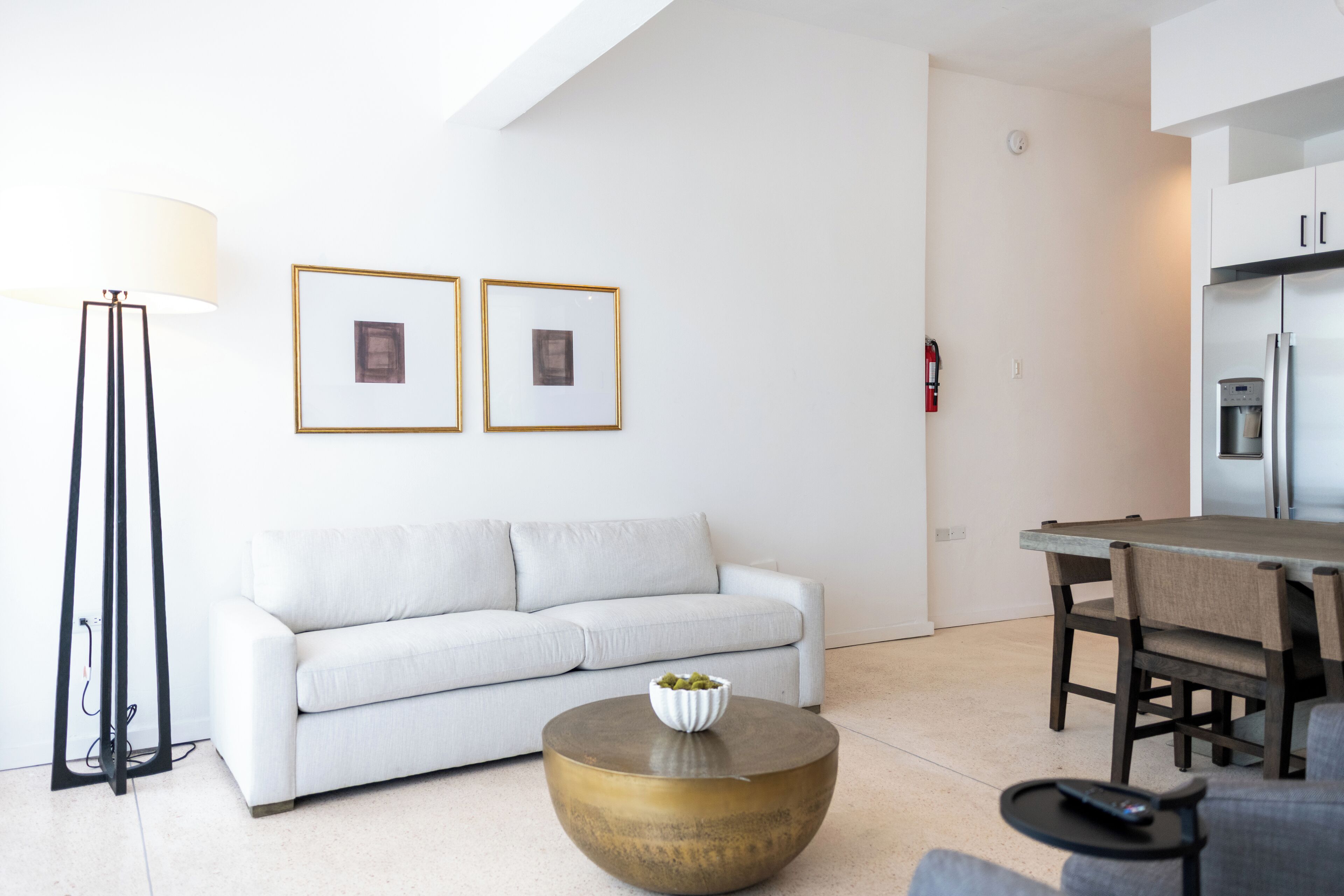 Een moderne woonkamer met een witte bank, een ronde salontafel met een schaal, een eettafel met stoelen en een keuken met een koelkast.