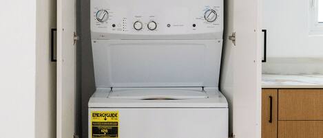 Un appareil compact combinant lave-linge et sèche-linge, doté d’un panneau de commande, d’une étiquette jaune indiquant les performances énergétiques et d’un meuble en bois situé à droite.