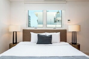 A hotel room with a bed, two bedside lamps, a window with a view of buildings, and a thermostat on the wall. - Ashford Apartments (San Juan)