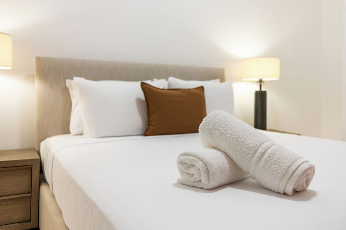 Een net opgemaakt bed met witte en bruine kussens, een opgerolde witte handdoek en een nachtkastje met een lamp.