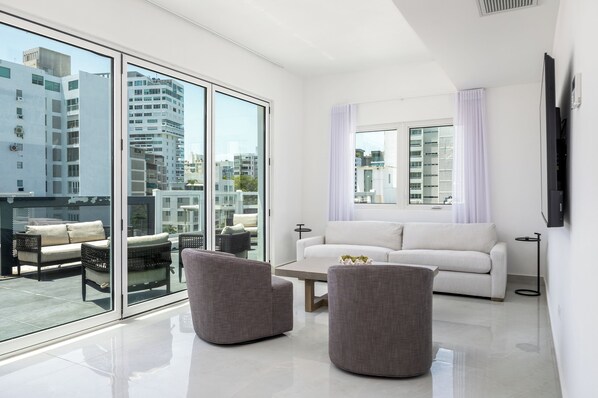A modern living room with a white sofa, two grey armchairs, a coffee table, and a large window with a view of city buildings. - Ashford Apartments (San Juan)