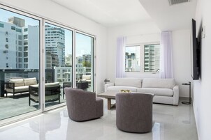 Una sala moderna con un sofá blanco, dos sillones grises, una mesa de centro y un ventanal con vistas a edificios de la ciudad.