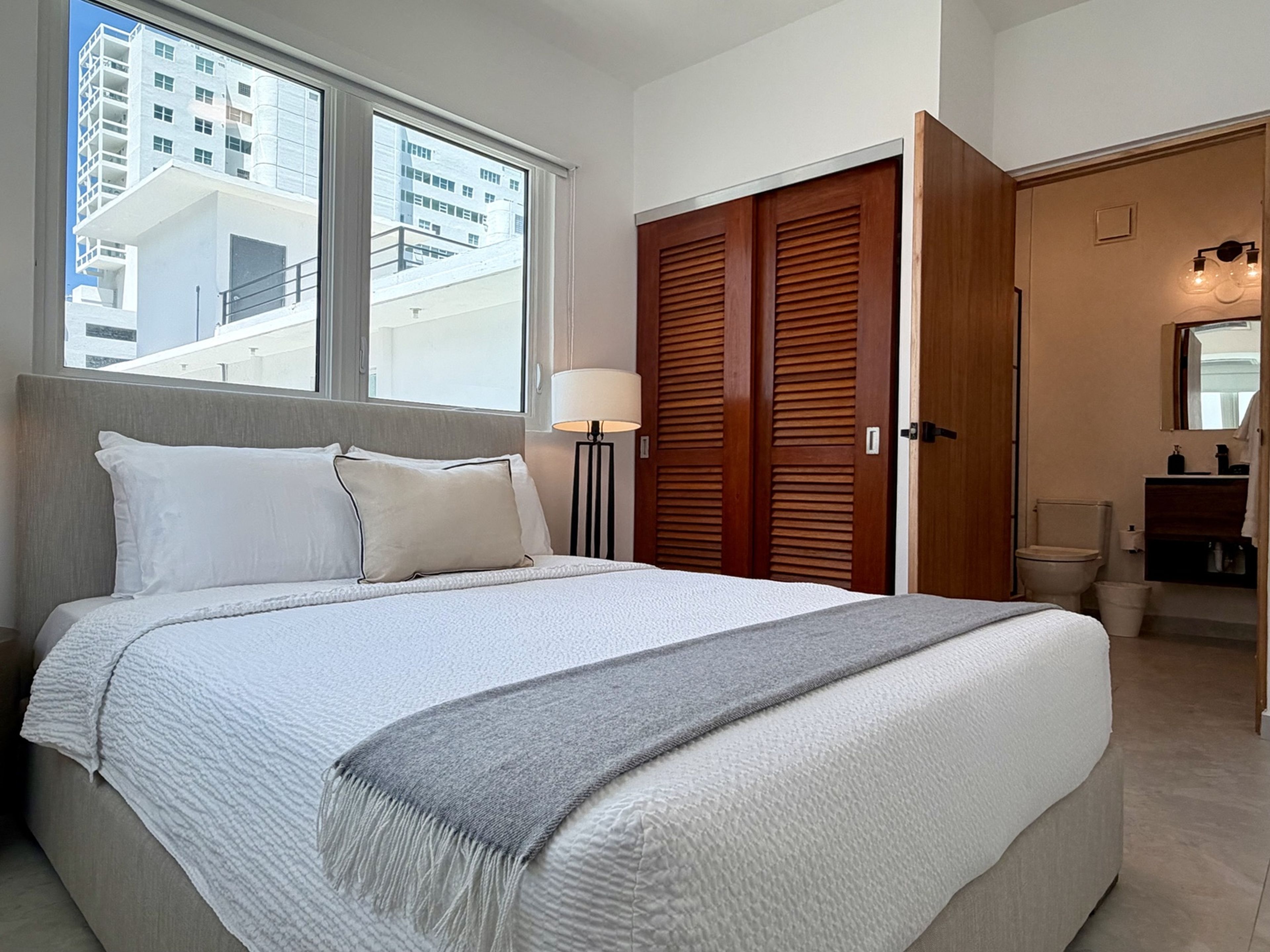 Une chambre d’hôtel avec un grand lit, une lampe de chevet, une armoire en bois et une vue sur le paysage urbain par la fenêtre.