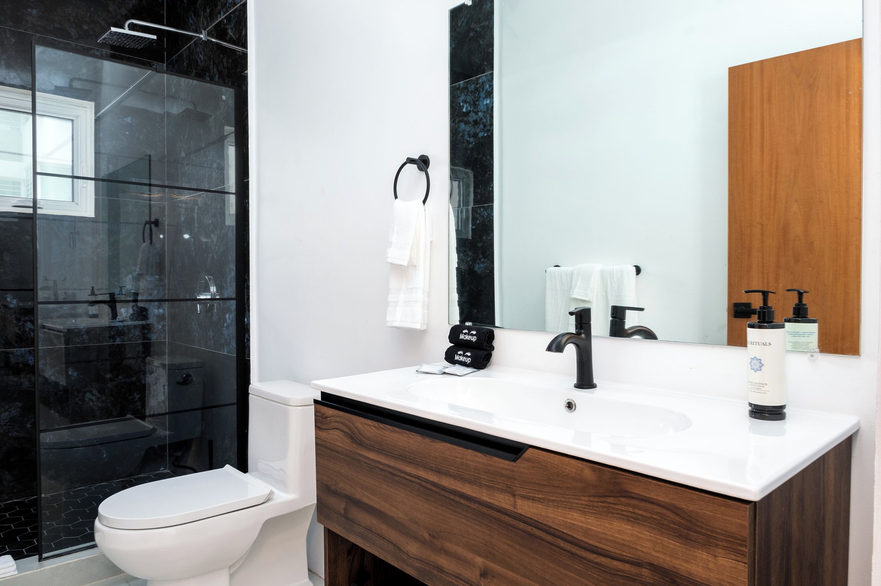 Un baño moderno con cabina de ducha de vidrio, inodoro blanco, tocador con gabinete de madera, espejo grande y grifo negro.