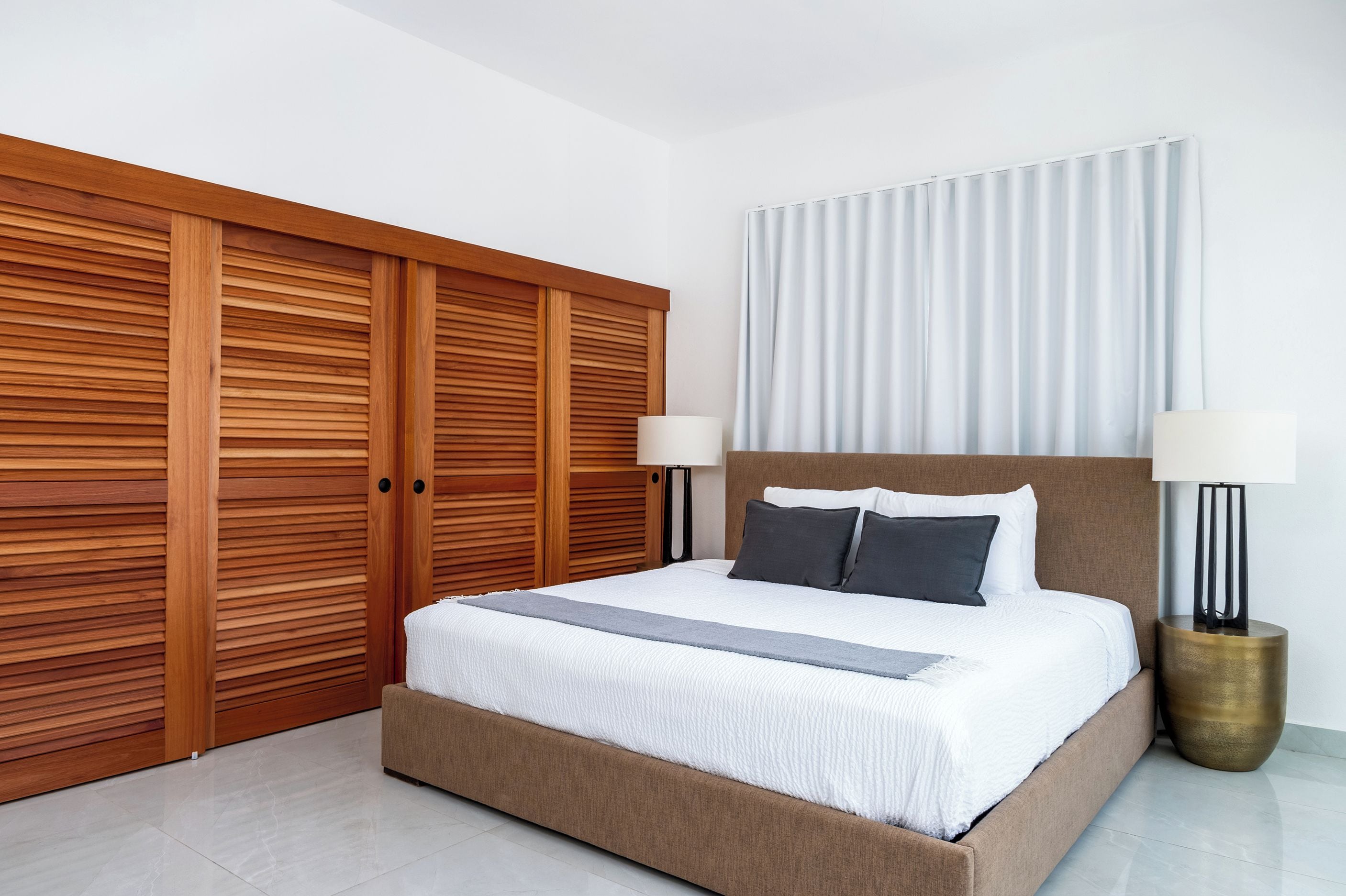 A bedroom with a large bed, two bedside lamps, a wooden wardrobe, and a window with curtains.