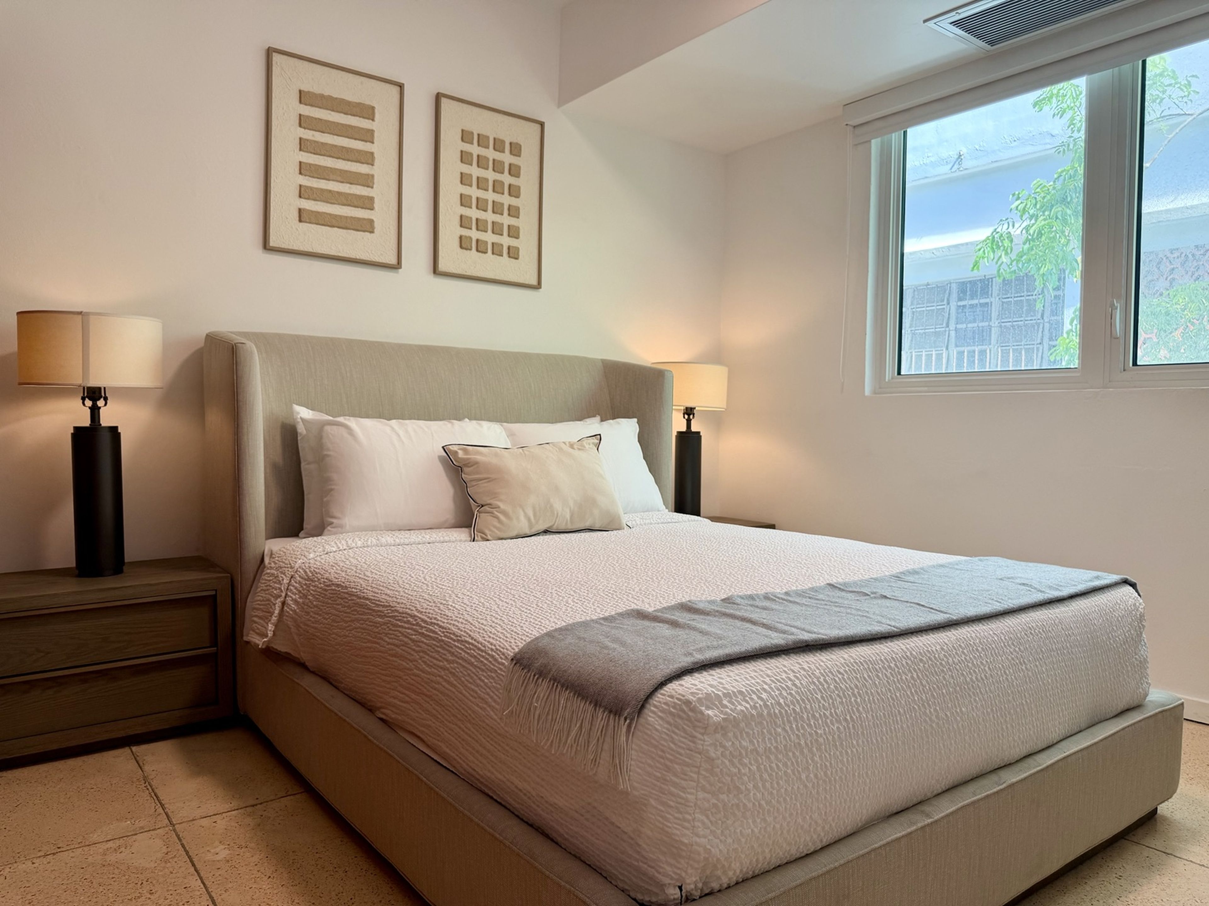 A neatly made bed with a headboard, two bedside lamps, and a window with a view of greenery.