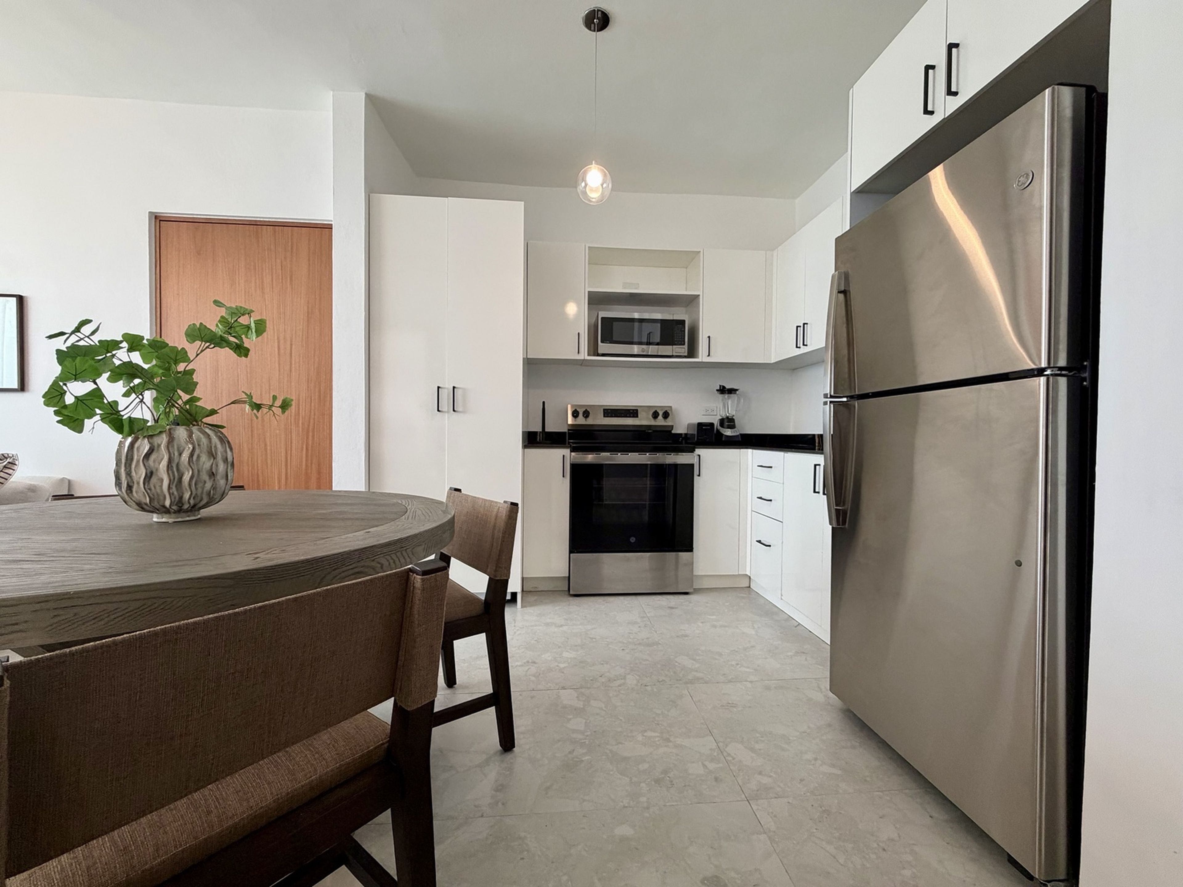 Cocina moderna con muebles blancos, un refrigerador de acero inoxidable, un microondas y un horno. Hay una mesa de comedor con sillas y una planta en maceta.