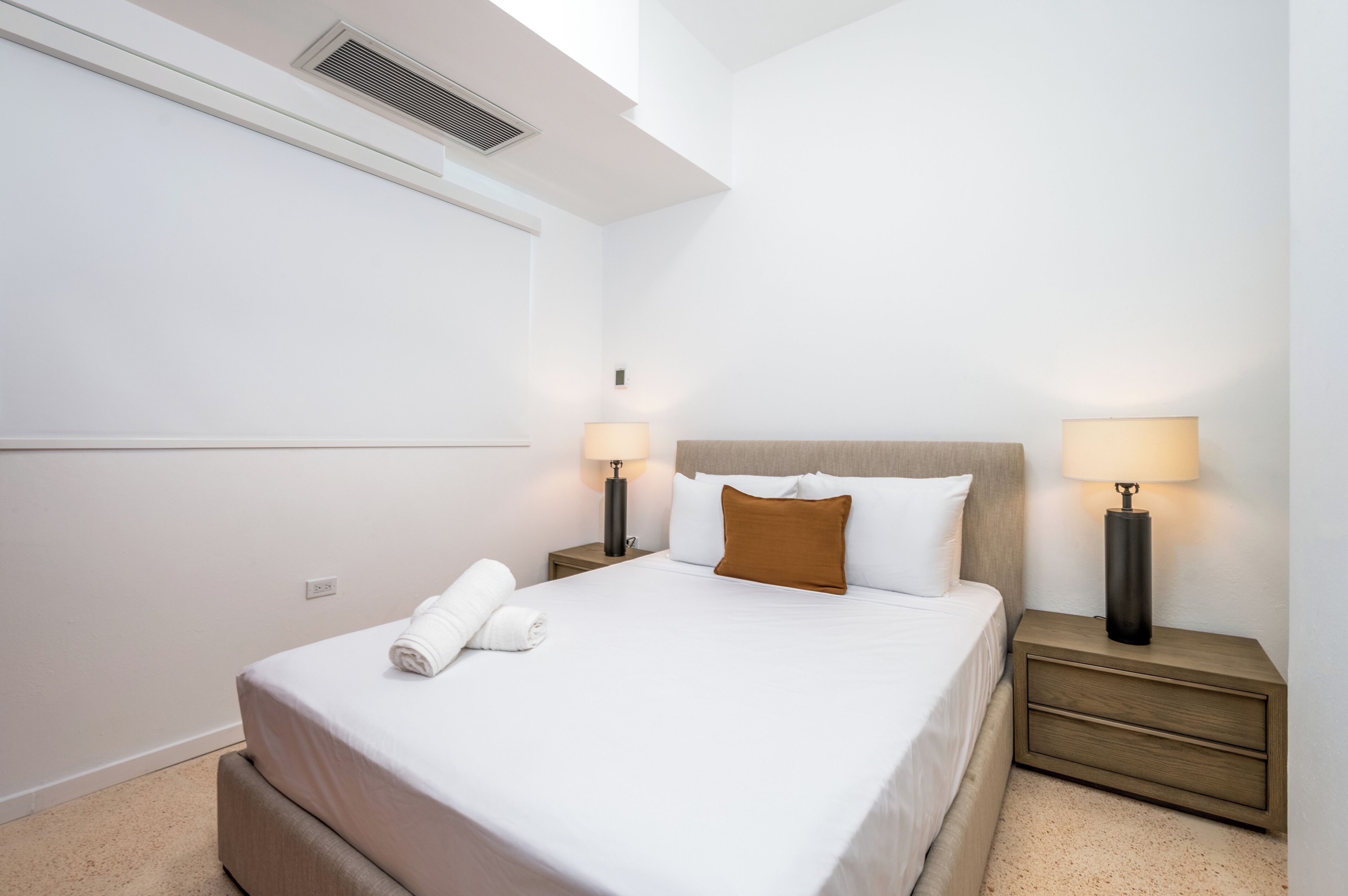 A hotel room with a bed, two bedside tables, and a wall-mounted air vent.