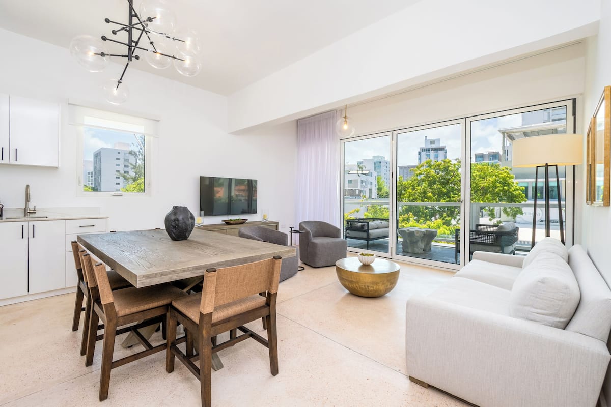 Een moderne woonkamer met een eethoek, grote ramen en een balkon met uitzicht op gebouwen en groen.