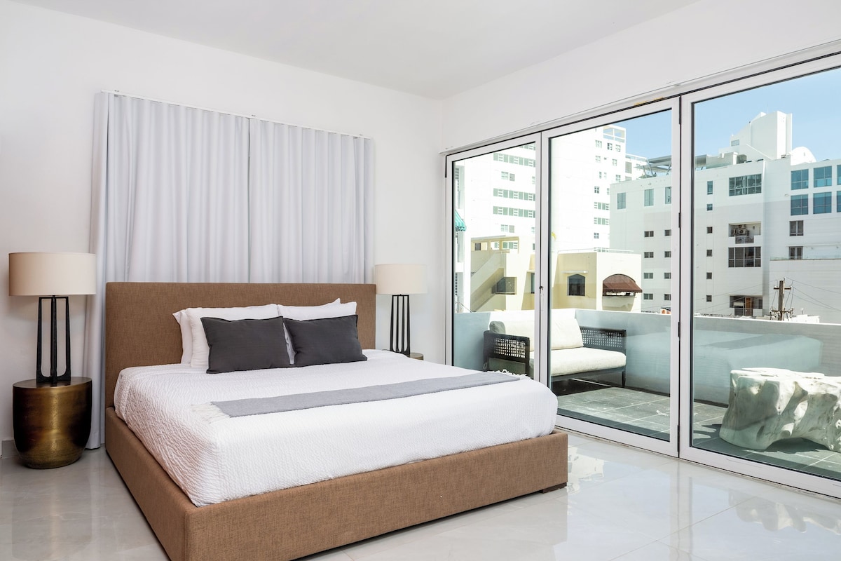 Una habitación de hotel moderna con una cama grande, vistas de la ciudad a través de ventanales del suelo al techo, y un balcón con mobiliario de exterior.
