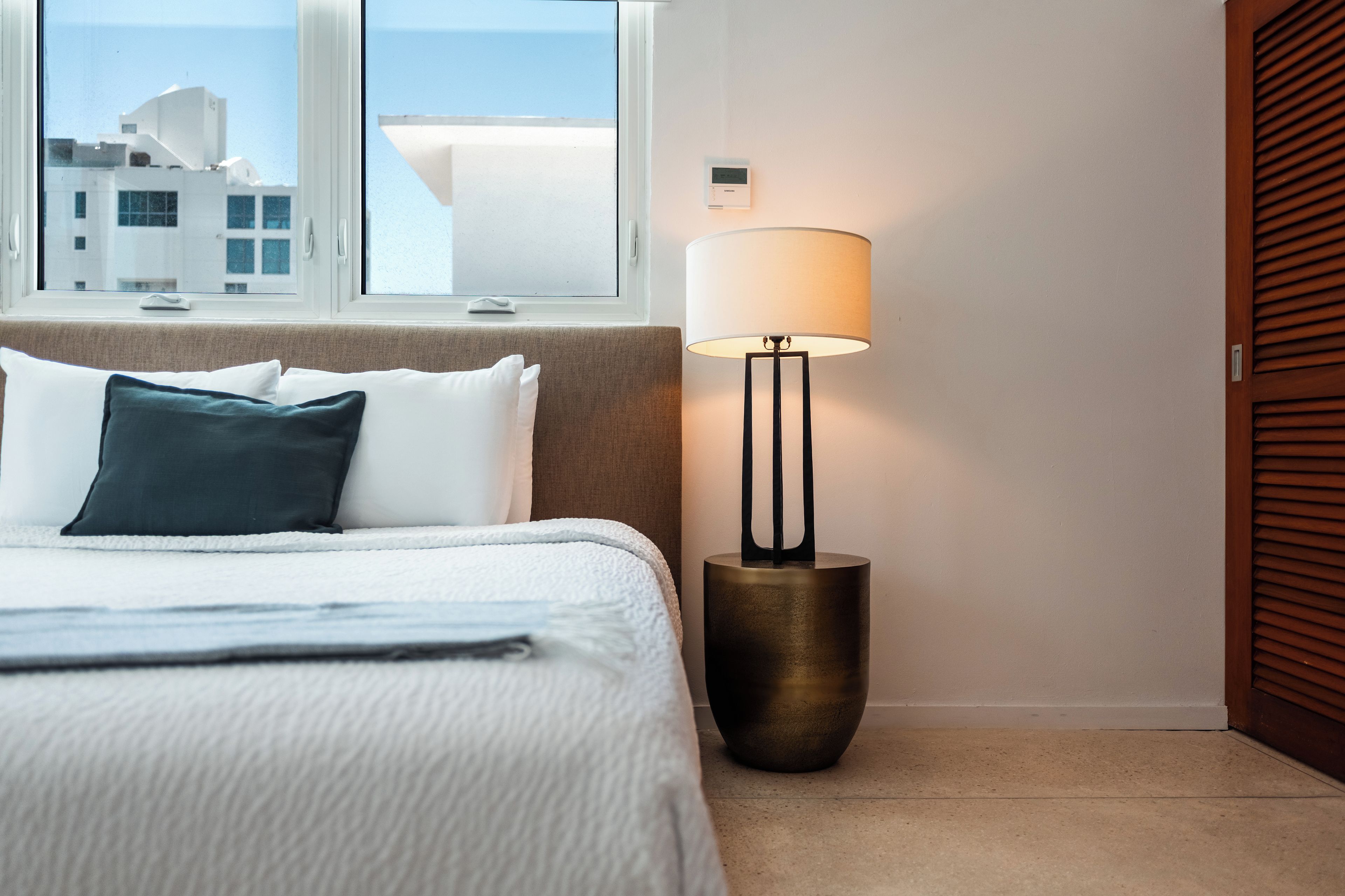 A hotel room with a bed, a nightstand, a lamp, and a view of buildings through the window.
