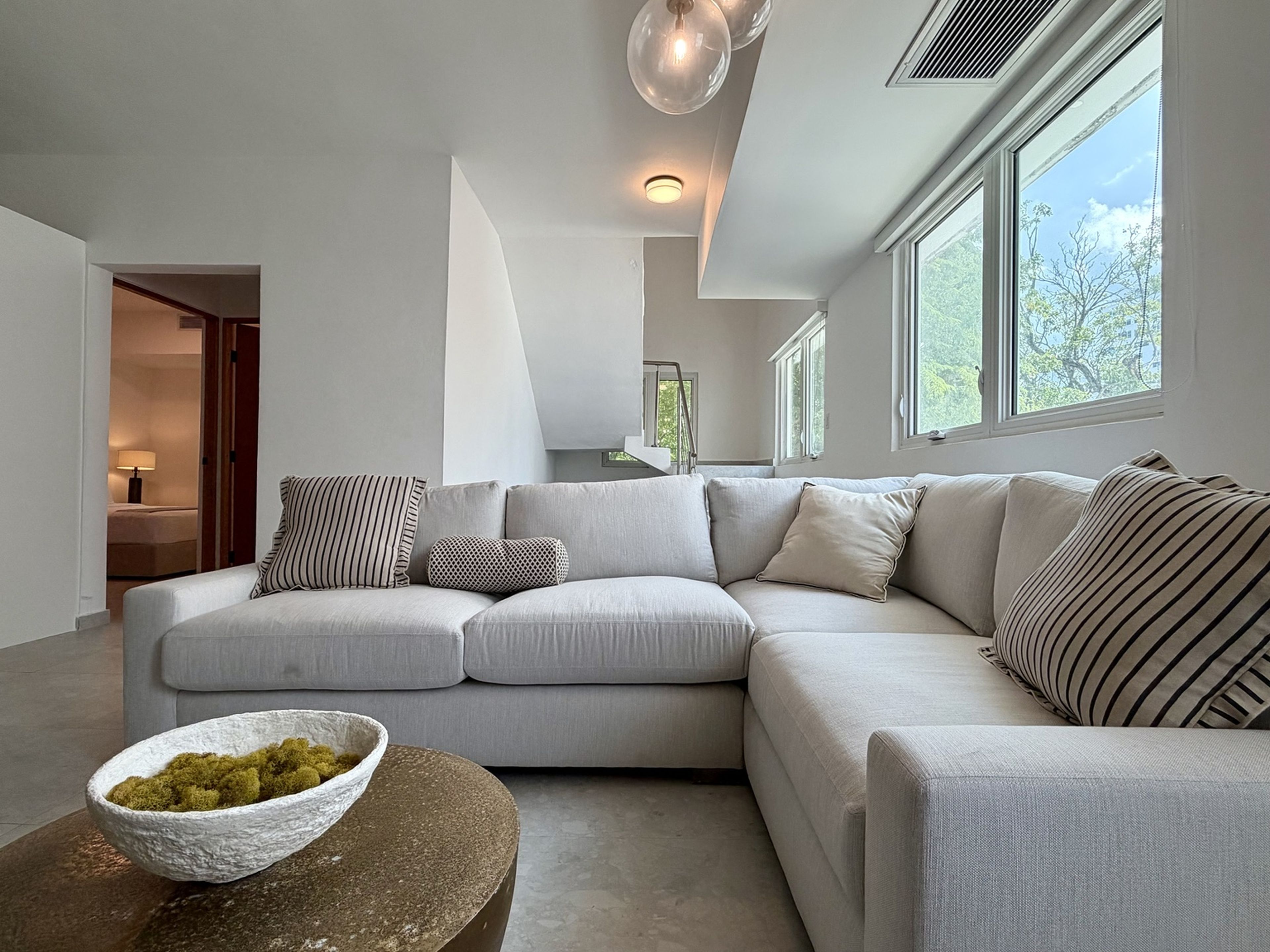 A modern living room with a grey sectional sofa, a round coffee table with a bowl of green plants, and large windows providing natural light.