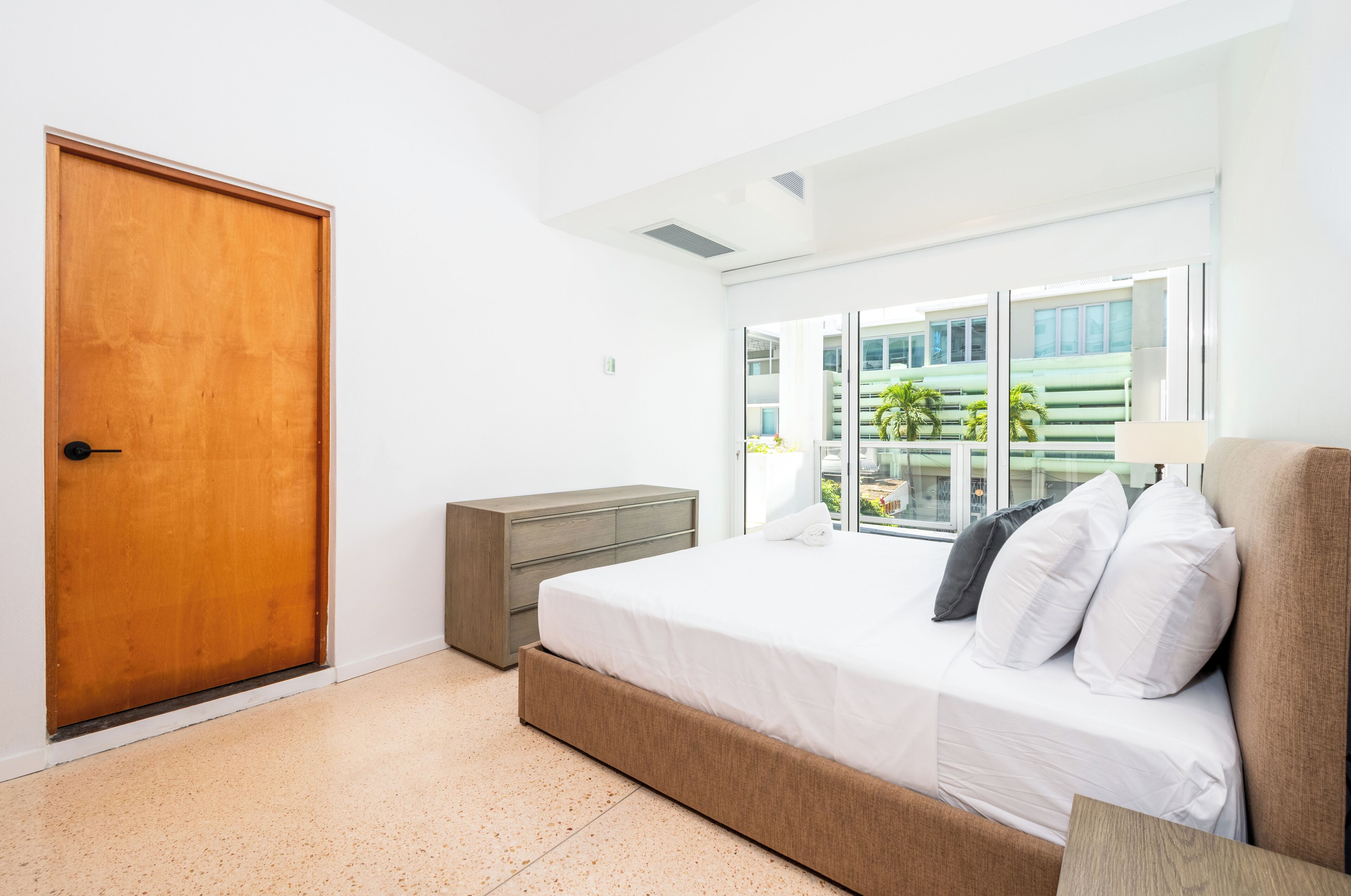 A bedroom with a wooden door, a dresser, a bed with white bedding, and a view of a building through large windows.