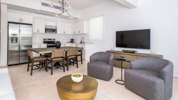 A modern kitchen with white cabinetry, stainless steel appliances, and a dining area with a wooden table and chairs. There is a large flat-screen TV mounted on the wall and a round coffee table in the living area.