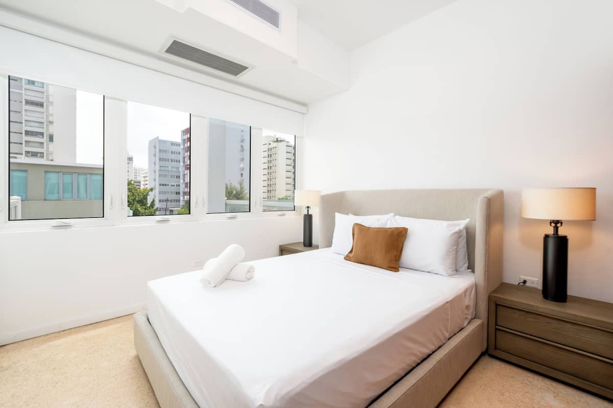 Een moderne hotelkamer met een groot bed, een nachtkastje, een lamp en uitzicht op gebouwen door grote ramen.