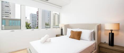 A modern hotel room with a large bed, a nightstand, a lamp, and a view of buildings through large windows.