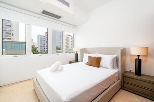 A modern hotel room with a large bed, a nightstand, a lamp, and a view of buildings through large windows.