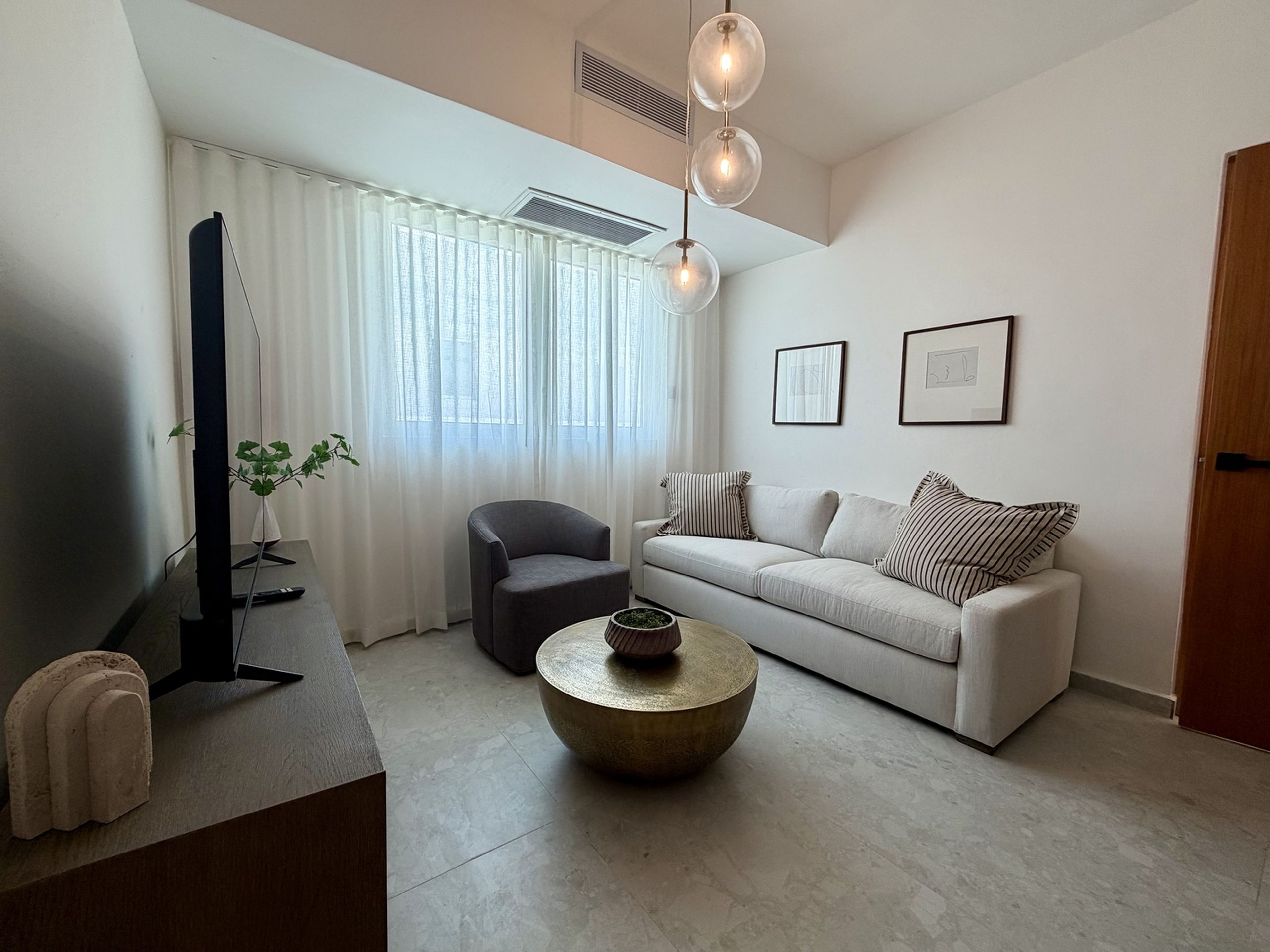 A modern living room with a flat-screen TV, a grey armchair, a white sofa with striped pillows, a round coffee table, and a large window with sheer curtains.