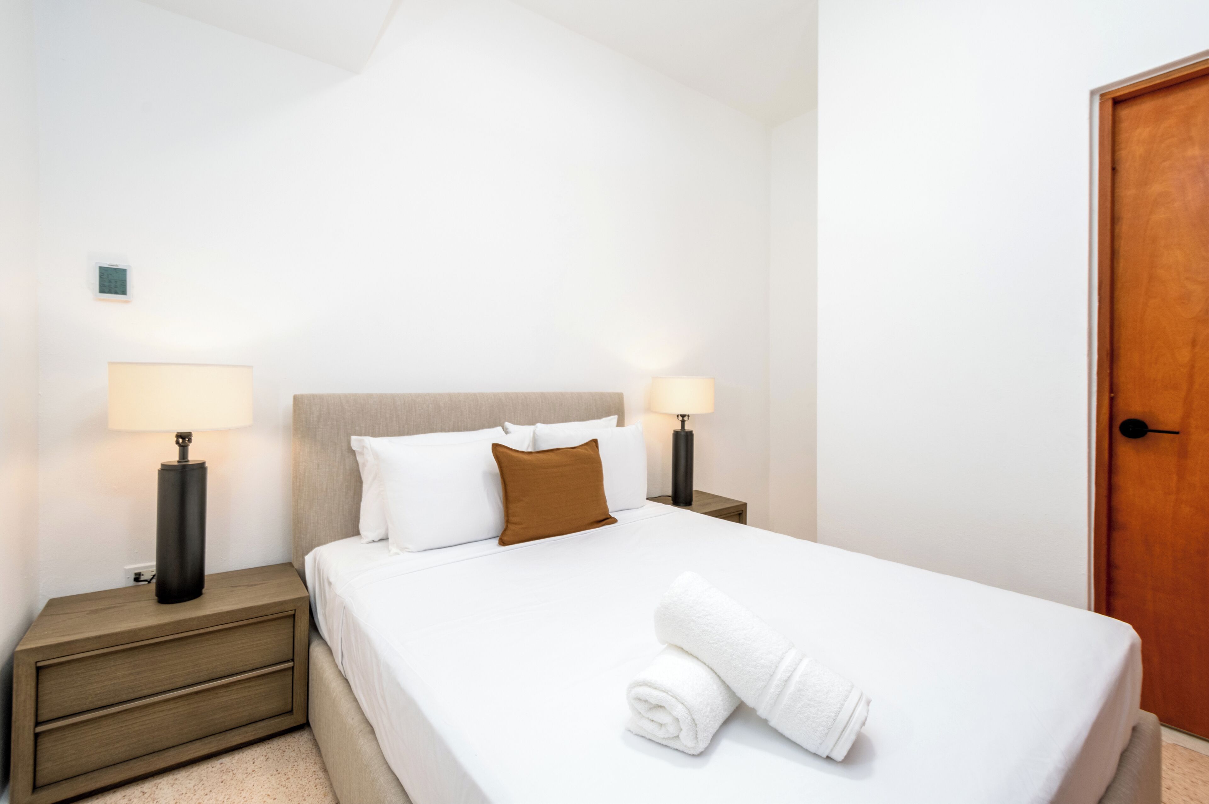 Een opgemaakt bed met witte lakens en een bruin kussen, twee bedlampen, een nachtkastje van hout met een lade, en een houten deur.