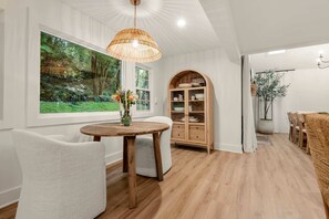 Un coin repas comprenant une table ronde en bois, des chaises à revêtement blanc, un buffet en bois doté de portes en verre et une grande fenêtre donnant sur la forêt.