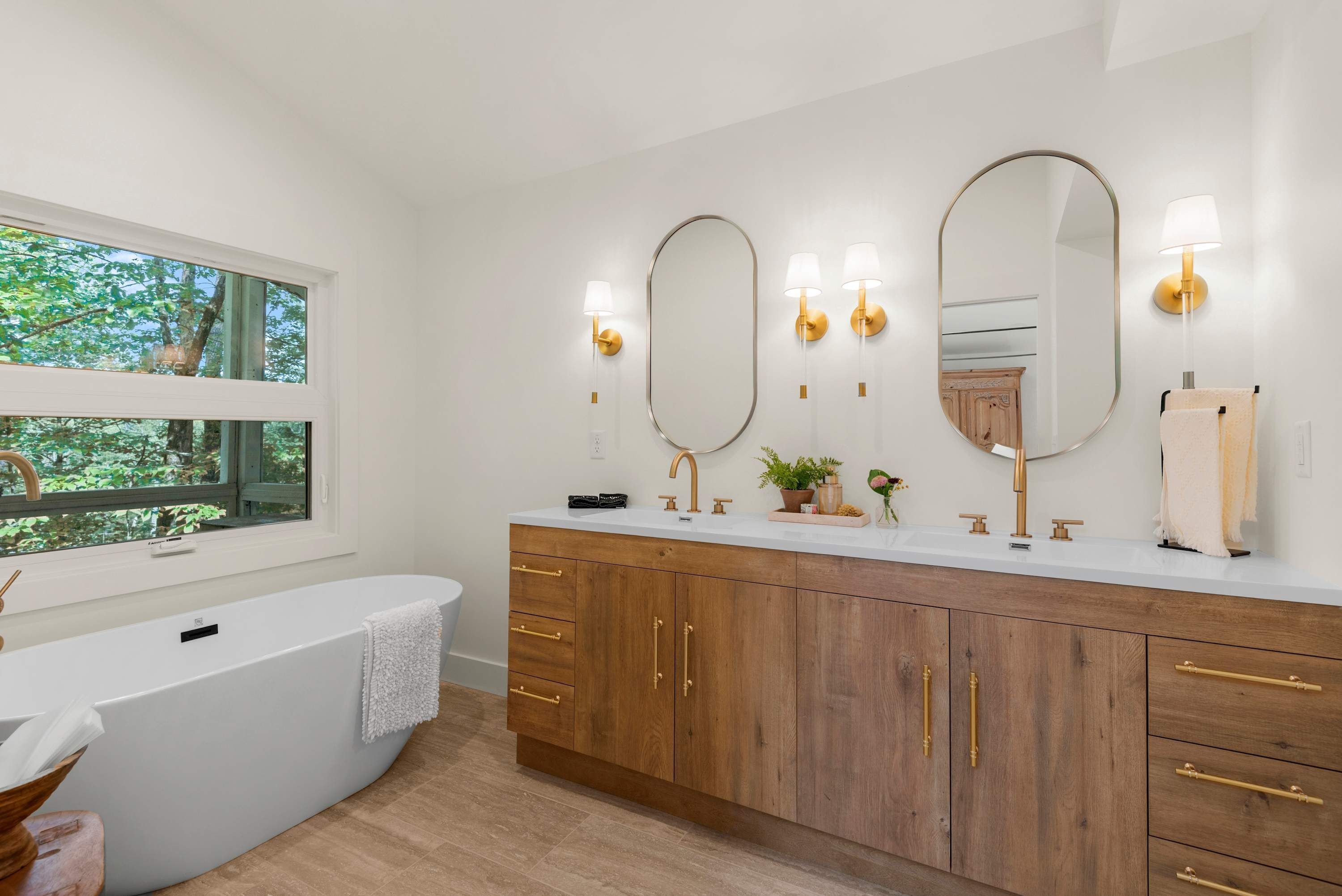 A modern bathroom with a freestanding bathtub, wooden vanity with gold handles, and three wall-mounted mirrors with lighting.