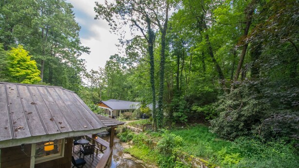 Une cabane en bois dotée d’une terrasse, entourée d’une forêt dense et traversée par un petit ruisseau.