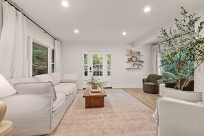Een moderne woonkamer met een beige vloerkleed, witte banken, een houten salontafel met boeken en een groot raam met gordijnen.