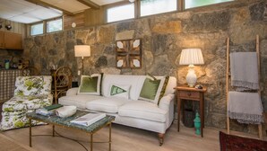 Un salon chaleureux avec un mur en pierre, un canapé blanc, un fauteuil à motifs floraux, une table basse en bois et une table d’appoint avec une lampe.