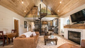 A spacious living room with a brown leather sofa, a coffee table, and a fireplace. A spiral staircase leads to an upper level. - Rustic-chic 2BR Cabin w Clawfoot Tub Near Trails (Hendersonville)