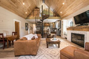 A spacious living room with a brown leather sofa, a coffee table, and a fireplace. A spiral staircase leads to an upper level. - Rustic-chic 2BR Cabin w Clawfoot Tub Near Trails (Hendersonville)