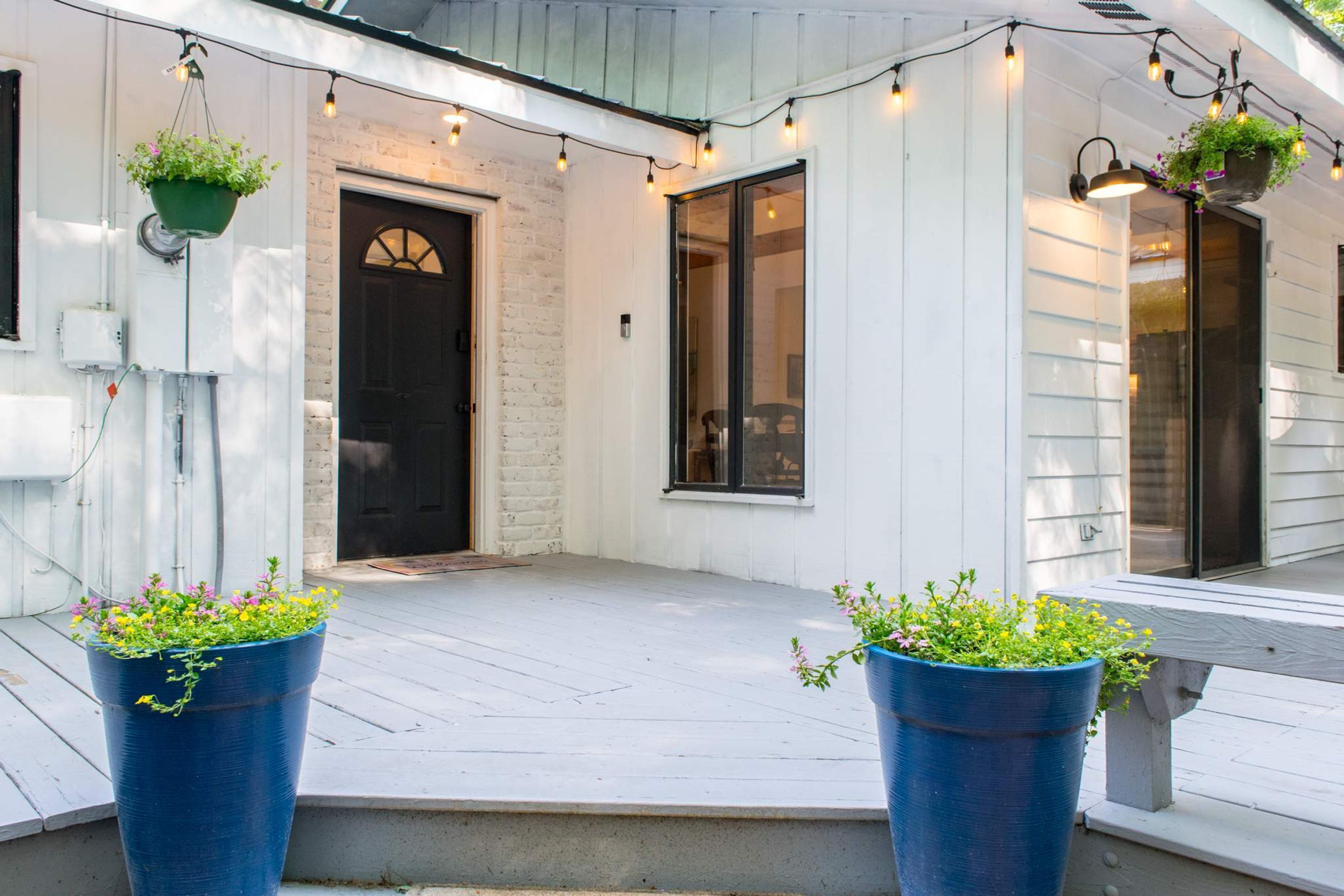 A porch with two large blue planters, a white wooden door, and string lights.