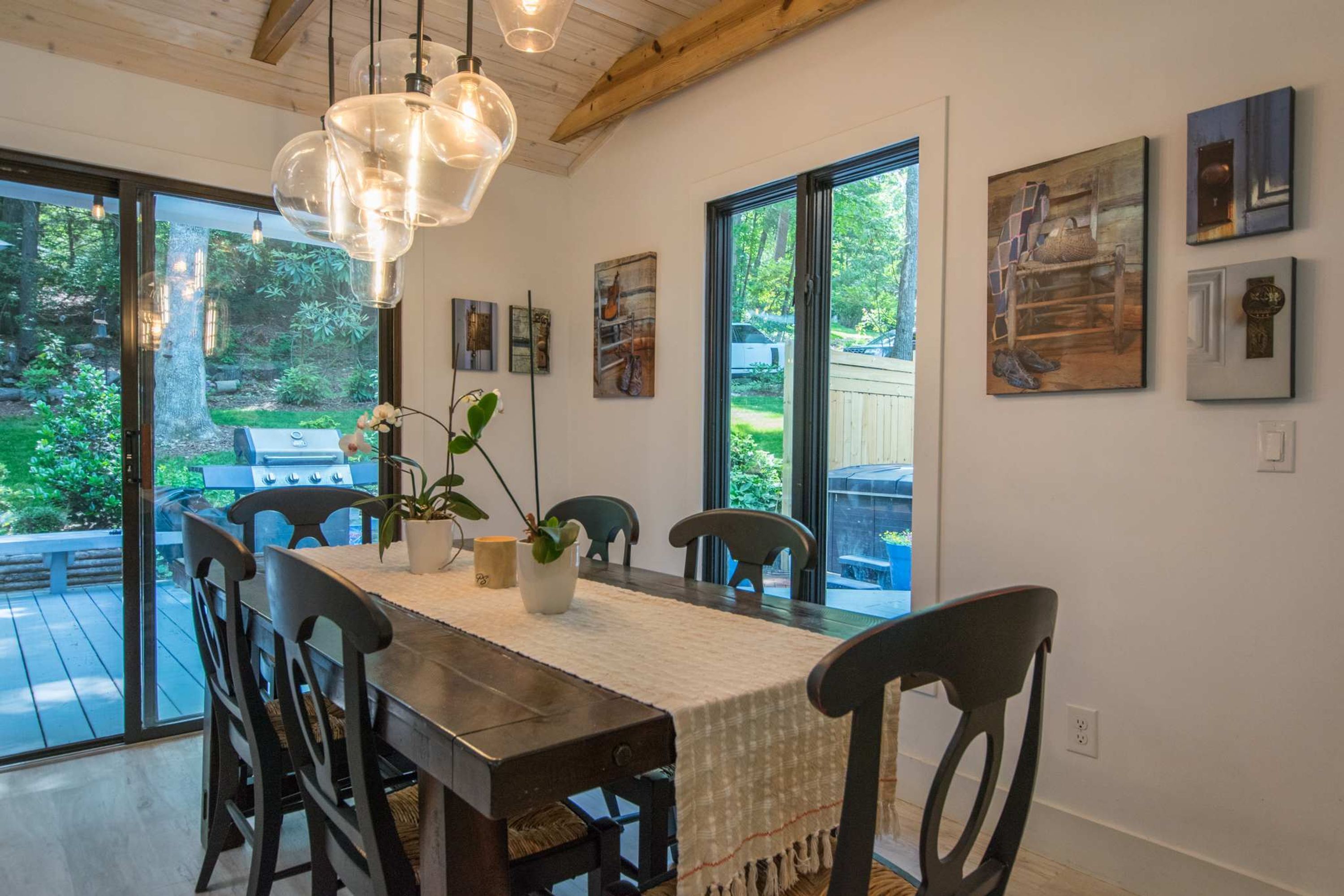 A dining area with a wooden table, chairs, and hanging pendant lights. There are framed artworks on the walls and a view of the outdoors through large windows.