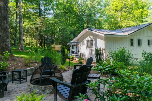 Une cabane dotée d’un foyer en pierre et d’un espace extérieur aménagé pour s’asseoir, le tout entouré d’une végétation luxuriante et d’arbres.