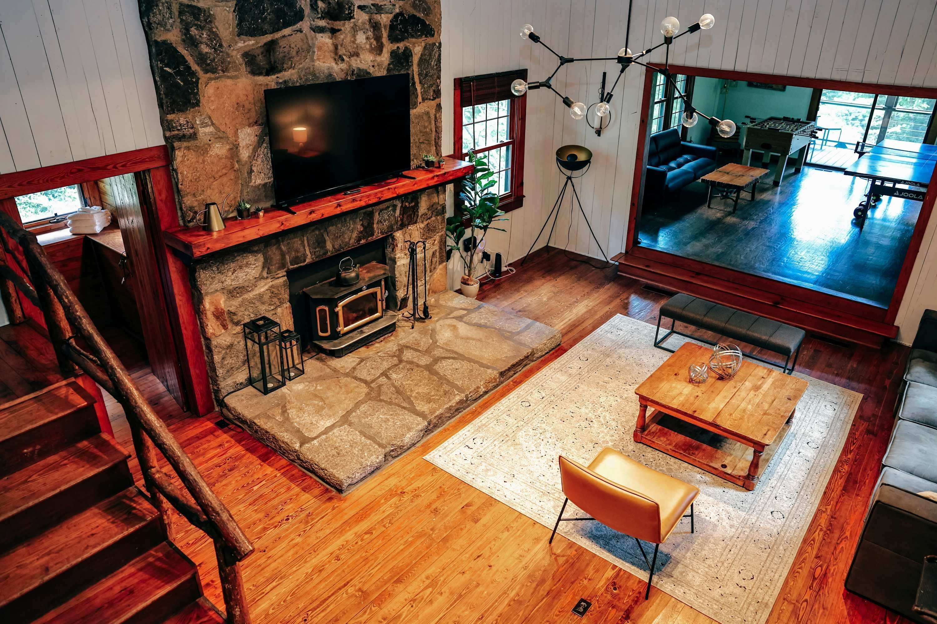 Un salon chaleureux avec une cheminée en pierre, un téléviseur à écran plat, un plancher en bois et une grande fenêtre donnant sur l’extérieur.