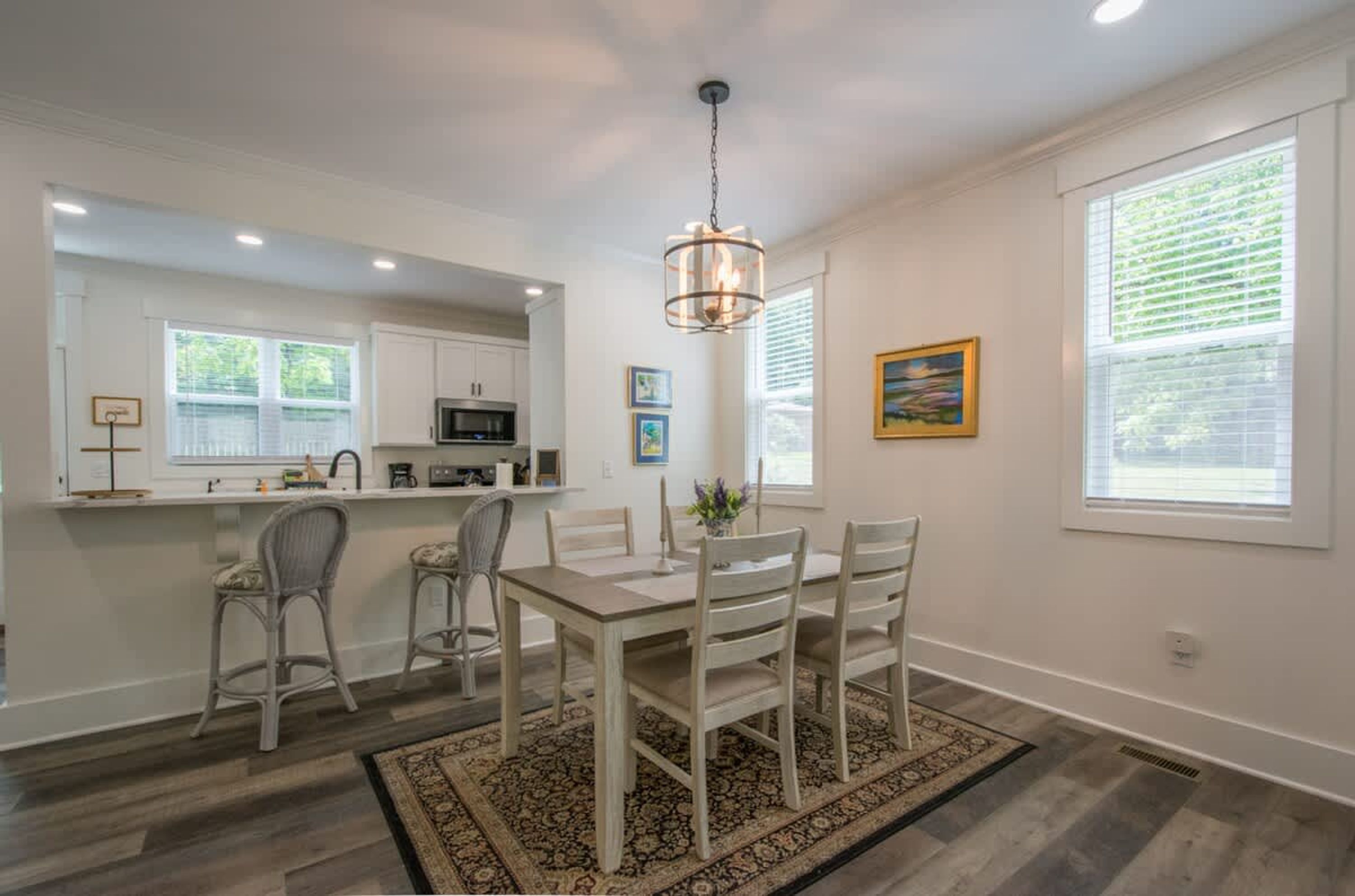 A dining area with a table and chairs, a kitchen in the background, and a large window with blinds.