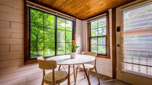 Une pièce avec un plafond en bois, de grandes fenêtres donnant sur un paysage verdoyant, une table ronde entourée de chaises et une plante en pot posée sur la table.