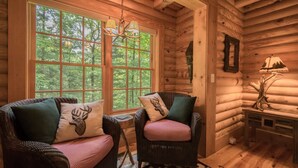 A log cabin interior with two wicker chairs, a chandelier, a large window with a view of trees, and a lamp on a wooden table. - Stylish 3BR Log Cabin w Fire Pit Near Trails (Zirconia)