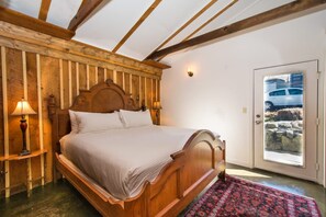 Ein hölzernes Bett in einem Raum mit schrägem Dach und einer Tür, durch die sich ein Auto und die Landschaft zeigen.