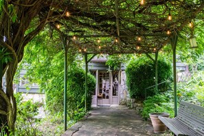 A covered garden pathway with a bench, surrounded by lush greenery and a building with a glass door. - 1BR Cottage w Patio Free Tix Near Downtown (Hendersonville)