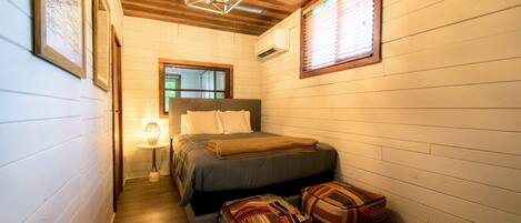 Un dormitorio acogedor con techo de madera, una cama con almohadas, una lámpara de noche y dos otomanas estampadas.