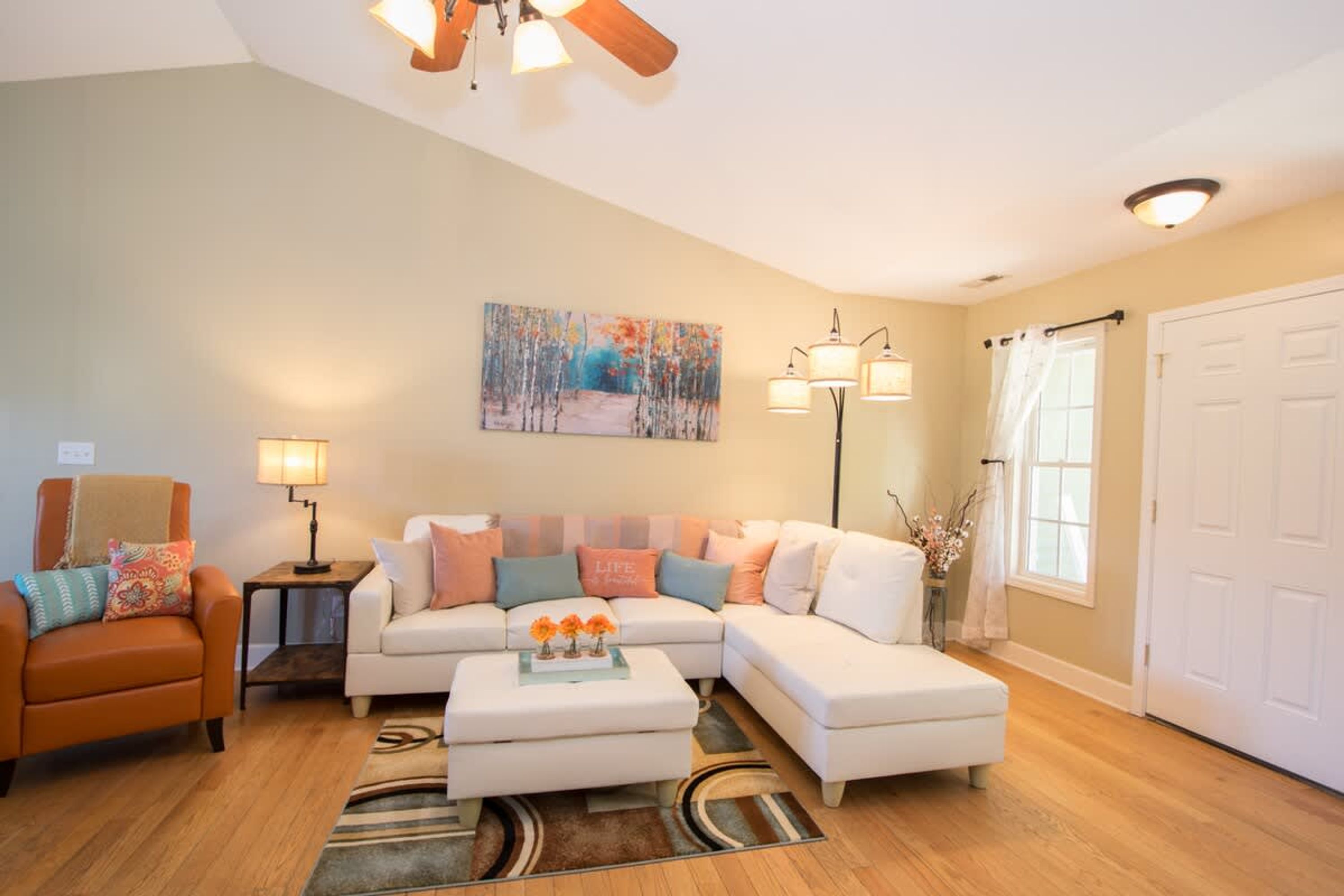 A living room with a sectional sofa, a small orange armchair, a coffee table, and a painting on the wall.