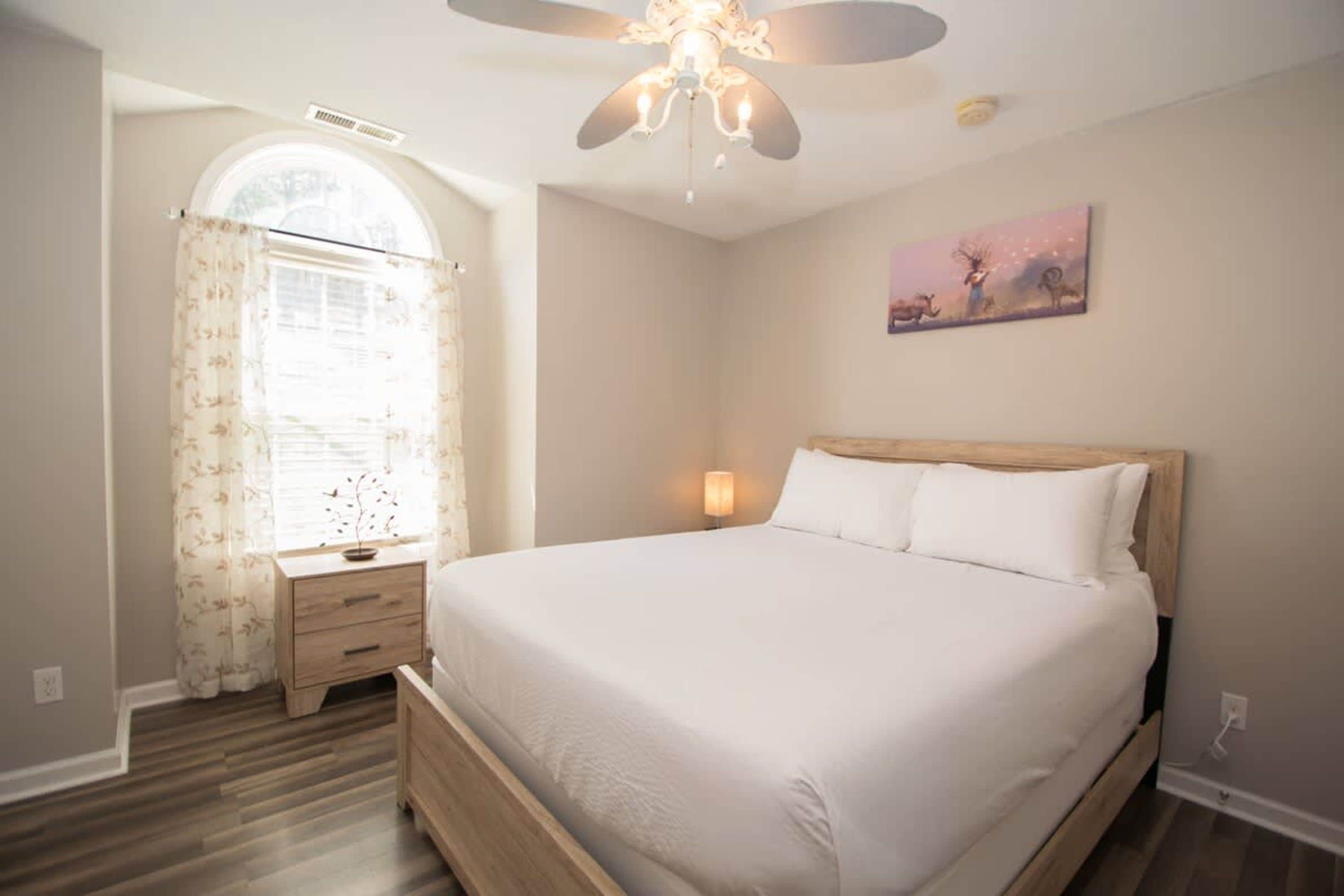 A bedroom with a bed, a nightstand, a lamp, a ceiling fan, and a window with curtains.