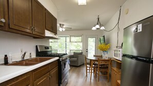 A kitchen with wooden cabinets, a stainless steel refrigerator, a stove, and a dining area with a table and chairs. - Lakeview 1BR Condo w Deck Walk to Village (Flat Rock)