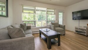 A living room with a sofa, armchairs, a coffee table, a TV, and a ceiling fan. - Lakeview 1BR Condo w Deck Walk to Village (Flat Rock)