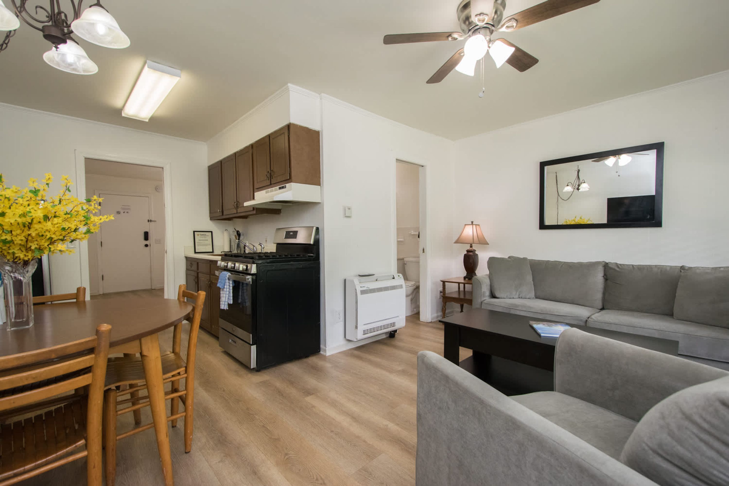 A modern kitchen with a stove, oven, and microwave, a dining area with a wooden table and chairs, a gray sofa, and a ceiling fan.