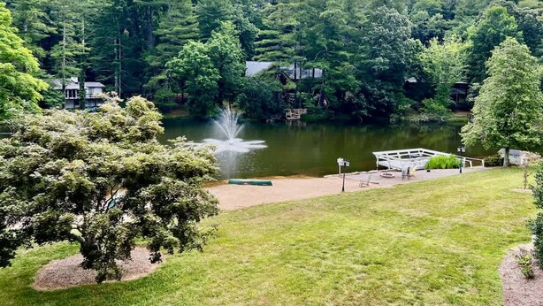 A serene lakeside setting with a fountain, surrounded by lush greenery and a dock. - Lakeview 1BR Condo w Deck Walk to Village (Flat Rock)