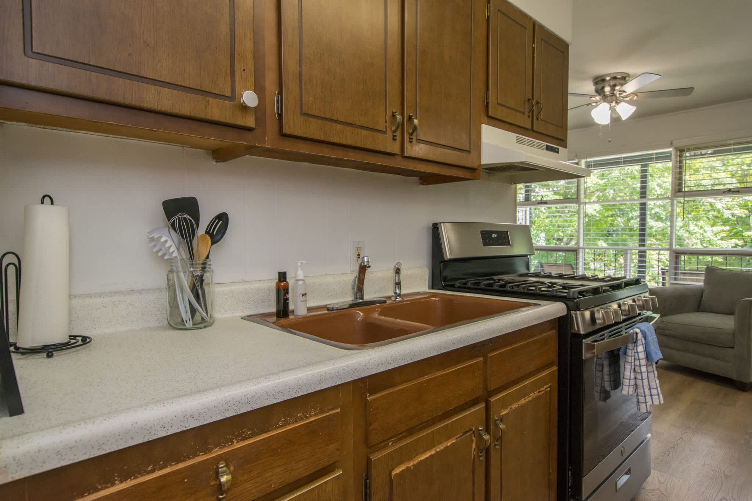 A kitchen with wooden cabinets, a stainless steel sink, a stove, and a window with a view of greenery.