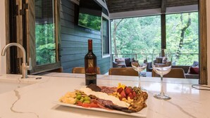 Une table à manger, agrémentée d’une planche de charcuterie, d’une bouteille de vin et de verres, est placée dans une pièce dotée de grandes fenêtres donnant sur une forêt.