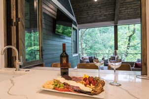 Una mesa de comedor con una tabla de embutidos, una botella de vino y copas, en un espacio con grandes ventanales con vista a un bosque.