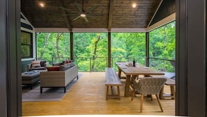 Un intérieur moderne avec un plafond en bois, de grandes fenêtres donnant sur la forêt, un coin repas avec une table et des chaises, et un espace détente avec un canapé et des coussins.